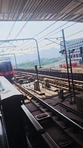 The video captures the arrival of a modern train at a station on a bright, sunny day. The shot is taken from a platform, looking across train tracks towards an approaching train. A metal railing is visible in the foreground, part of the platform's edge.

The train has a sleek design with a red front, which features an illuminated display showing Chinese characters, likely indicating its route or destination. As the train moves past the camera, its side reveals a grey metallic finish. On one of the train cars, the number "010542" is clearly visible.

Overhead, a complex network of electrical lines and support structures for the train's power system spans the tracks, partially covered by the station's roof. In the background, beyond the railway lines, several modern buildings with repetitive window patterns are visible, suggesting an urban or industrial area. Further in the distance, hills or mountains can be seen on the horizon. The train appears to be slowing down, indicating its arrival at the platform in what is identified as Chongqing High-tech Zone, China.
