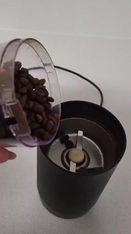 Here is a description of the video.

The video shows a person pouring coffee beans into a black electric coffee grinder on a white countertop. The person then puts the lid on the grinder.