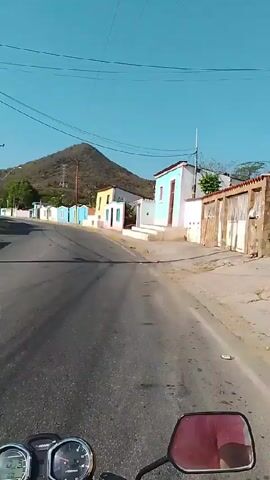 A motorcyclist is riding down a street lined with colorful houses. The houses are painted in a variety of bright colors, including blue, pink, and yellow. A mountain can be seen in the distance, under a clear blue sky. The road is paved and there are power lines running overhead. The camera is positioned on the motorcycle, providing a first-person perspective of the ride. The motorcycle is moving at a moderate speed, and the sound of the engine can be heard.
