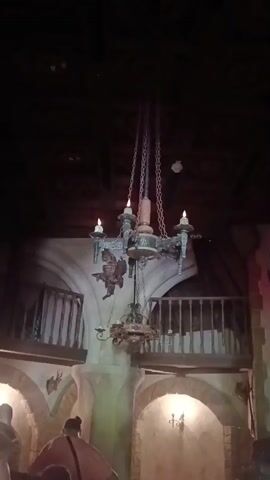 The video shows an interior scene with a large, rustic chandelier hanging from the ceiling. The chandelier has candle-like lights and decorative elements, including what appears to be a gargoyle figure. Below the chandelier, there is a balcony with wooden railings. The walls are painted in earthy tones and feature architectural elements like arched doorways and textured plaster. On the walls, there are mounted deer antlers and crossed axes, suggesting a medieval or rustic theme. A fireplace is visible on the right side of the frame, adorned with more antlers and decorative objects. Several people are present, partially visible in the foreground, indicating that this is likely a public space like a restaurant or attraction. The lighting is dim, creating a somewhat dramatic atmosphere.