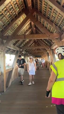 The video shows a perspective from behind a person wearing a bright yellow safety vest and a white helmet, walking along a wooden covered bridge. The bridge has a visible wooden truss roof structure above. Several other people are walking in the same direction as the viewer. To the immediate left of the viewer, a man wearing a black t-shirt and khaki shorts walks by. Further down the bridge, a woman in a white striped t-shirt and a white skirt walks, followed by other individuals and couples. One man is carrying a baby in a carrier on his chest. The bridge appears to be a tourist attraction, with people casually walking through it. The lighting suggests it is daytime with sunlight filtering through the open sides of the bridge.
