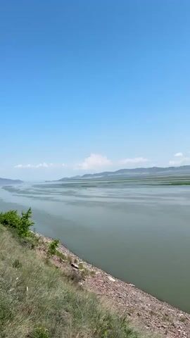 A wide body of water stretches towards a horizon lined with green plains and rolling hills under a bright blue sky dotted with white clouds. The foreground shows a gravelly bank with dry grass. A group of people, some carrying backpacks, are walking along a dirt path on a grassy incline to the right of the water. One person in the foreground, wearing a pink baseball cap and white shirt, is facing away from the camera, looking towards the water and the group.