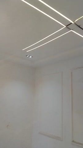 The video shows an interior scene with a white ceiling and walls. A modern chandelier with a circular base and a curved arm holding three spherical bulbs hangs from the ceiling. There are also two linear LED lights embedded in the ceiling, one on the left side and one on the right side, which appear to be recessed. In the upper right corner of the ceiling, there are two small, square-shaped spotlights. The walls are plain and white, with a dark square electrical box visible on the wall in the lower left portion of the frame. The floor is partially visible at the bottom of the frame and appears to be made of light-colored tiles with dark grout lines.
