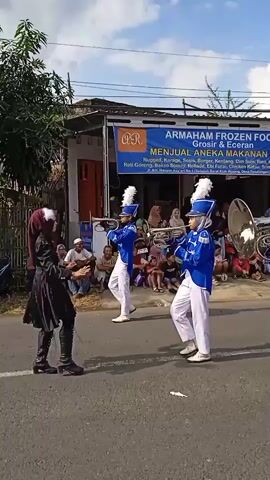The video shows a marching band performing in the street. The band members are wearing blue and white uniforms with tall white plumes on their hats. They are playing silver horns and walking in formation. A woman in a black coat and hijab is walking alongside the band. Spectators sit on the side of the road to watch the performance. A store with a sign that says “Armaham Frozen Food” is in the background. The sky is partly cloudy.