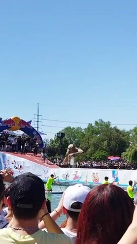 The video shows a Red Bull Flugtag event with crowds watching as a participant rides a bicycle off a ramp and into the water. The participant is riding a bike down the ramp. There are people in the water with what looks like canoes. There are many spectators and the weather is sunny. Some of the spectators are recording with their phones.