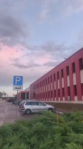 The video shows an exterior view of a modern industrial or commercial building with a red facade and white trim. The building has multiple windows and air conditioning units visible. In the foreground, there is a parking lot with several cars parked, including a silver station wagon. A blue parking sign is visible. Lush green bushes and grass are in the immediate foreground. The sky is visible, with clouds displaying shades of pink and purple, suggesting either sunrise or sunset. Streetlights are present along the road. In the distance, more buildings and trees can be seen. A pedestrian crossing is visible, and a person wearing a backpack and dark clothing is crossing the street. The ground appears wet, possibly from recent rain. The overall scene suggests an urban or suburban environment during twilight.