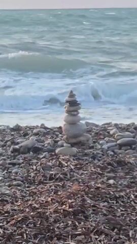 A rock cairn stands on a rocky beach in front of the ocean. The ocean has waves and the sky is light. The beach is covered in small stones and dried seaweed.