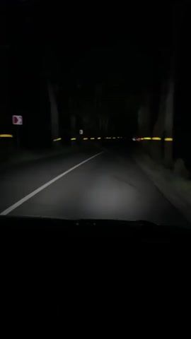 The video shows a point-of-view shot from inside a car driving on a dark road at night. The headlights illuminate the asphalt road with a white dividing line. Yellow markings are visible on the trees lining the road. In the distance, the red taillights of another vehicle can be seen. The surrounding environment is dark, suggesting it is nighttime in a wooded or rural area. The camera is mounted inside the car, looking forward through the windshield.