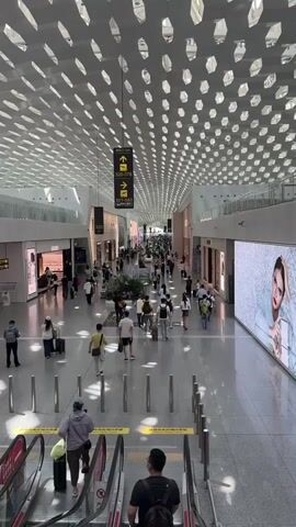 The video shows a wide, modern airport terminal with a high, intricately patterned ceiling. Many people are walking in different directions along a large, open concourse. Some are pulling luggage, indicating travel. Stores are visible on either side of the concourse, suggesting a commercial area within the airport. Signage with arrows points to different gates or areas, such as "320-378" and "501-583". Escalators are also visible, with some individuals descending them. A large digital screen displaying an advertisement is mounted on the right side of the concourse. The overall environment appears clean, spacious, and well-lit, characteristic of a contemporary international airport.