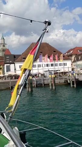 The video shows a view from a boat moving through green water, leaving a foamy wake. In the background, there is a waterfront town with buildings and a pier. A German flag is prominently displayed on a pole on the boat. Several other flags are visible on buildings in the town. The sky is partly cloudy. There are people visible on the pier and in the background. The scene appears to be a sunny day in a European town.