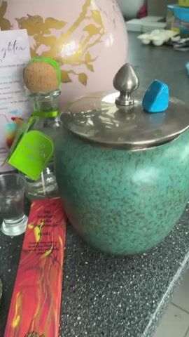 The camera pans slowly across a cluttered countertop, revealing a collection of objects. These include a silver incense holder with a meditating Buddha figure and seven colorful stones running down its length, a shot glass filled with clear liquid, a bottle of Patron tequila with a green tag, a box of “The Mother’s India Fragrances” incense, a turquoise ceramic container with a silver lid, a pink vase with gold floral designs, and a greeting card for a “Wonderful Daughter.” A wine glass filled with a clear liquid is also visible in the background. The countertop appears to be a dark, speckled material. The camera movement focuses on showcasing the various items on the counter.