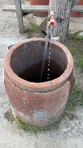 Here’s a concise description of the video:
The video shows an old, cylindrical ceramic container with a wide opening, with water dripping into it from a pipe. The container has a weathered, reddish-brown exterior with signs of age and wear, set on a concrete surface with patches of grass. In the background, a wooden structure provides shade, under which several similar ceramic containers are stacked. A tree trunk stands beside the container, and a sign with text and a phone number is visible on the wooden structure. The camera slowly zooms out, revealing more of the outdoor setting.