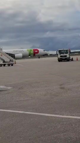 The video shows an airplane on a tarmac, bearing the "Portugal" name and logo. Adjacent to the plane is a set of passenger stairs, a white truck, and a yellow and white vehicle labeled "ELECTRIC". Orange cones are also visible on the tarmac near the truck. The sky is overcast with heavy clouds. The video is shot from a static viewpoint with a minimal amount of camera movement. There are no people in the frame.