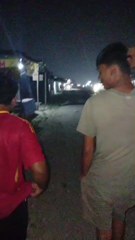 Two people are walking away from the camera at night. The person in the foreground is wearing a red shirt with yellow accents and black shorts. The person behind them is wearing a light-colored t-shirt and shorts. They are walking down a dirt path next to some buildings. There are lights visible in the distance. One of the buildings has a yellow sign with red writing.