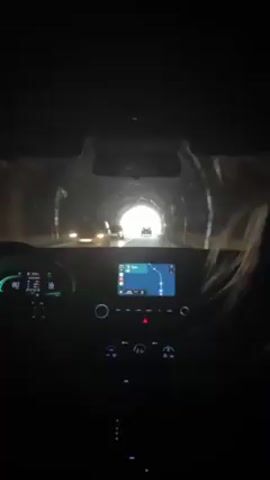 The video is taken from inside a car driving through a tunnel and out into a landscape with hills and other cars. The dashboard shows the car's speed and navigation. A person is driving, and there is a passenger beside them. The car exits the tunnel into daylight.