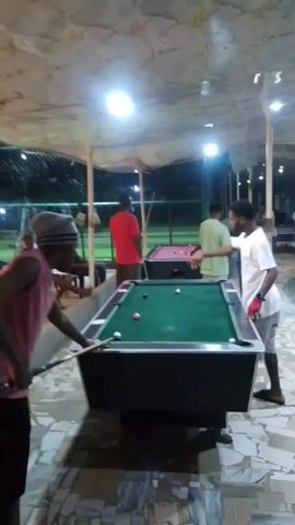 The video shows a group of people playing pool outdoors under a covered area. There are at least three pool tables visible, with people gathered around each. One man, wearing a pink sleeveless shirt and a gray hat, is preparing to take a shot on a green pool table. Another man in a white shirt stands nearby, watching. In the background, another person in a red shirt is playing on a red pool table. The area is lit by overhead lights. The floor is made of patterned tiles. The surroundings appear to be a recreational area or a casual outdoor space.
