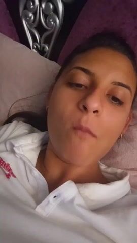 A close-up, eye-level, selfie-style video shows a young woman lying down and looking into the camera. She is wearing a white polo shirt. Her hair is dark and pulled back. The background appears to be a bed with pillows and a headboard. The lighting is dim.