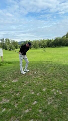 A man in a black jacket and light gray pants is playing golf on a green course. He swings a golf club and hits a white golf ball. In the background, there is a golf course with trees and a cloudy sky.