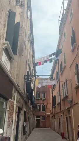 The video captures a narrow alleyway in Venice, Italy, flanked by tall, aged buildings. The building on the left is a lighter beige, while the one on the right has a reddish-brown facade. Both buildings feature multiple windows, some equipped with dark shutters. Several lines of laundry, consisting of various colorful shirts and t-shirts, are strung across the alley between the buildings, hanging to dry under an overcast sky. A white street sign with black lettering, reading "CALLE LARGA," is visible on the left building. In the opening seconds, a woman with dark hair, wearing a white top, is seen smiling in the bottom right corner of the frame. The camera performs a slight zoom, bringing the hanging laundry into sharper focus. The scene depicts a common sight of daily life in Venice.