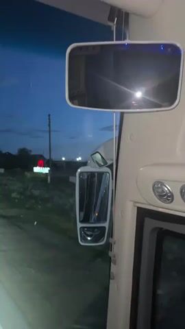 This media file captures a view from inside a vehicle, likely a bus or truck, as it travels along a road at dusk. The exterior mirrors are visible, reflecting the dim light and the road ahead. The scene outside depicts a rural or suburban setting in Baiqadam, Kazakhstan. Houses and trees line the road, with streetlights beginning to illuminate the darkening sky. A red neon sign, possibly for a business, is visible in the distance. Traffic signals and road signs, including one indicating a speed limit of 20, are also present. The sky is a deep blue, transitioning into twilight, with some clouds and a bright light, possibly the moon or a streetlamp, visible. The headlights of the vehicle are on, casting a bright path on the wet asphalt road. A partial view of a license plate with Cyrillic text, "АКБА", is visible at the bottom of the frame. The overall atmosphere is calm and transitional, marking the end of the day.