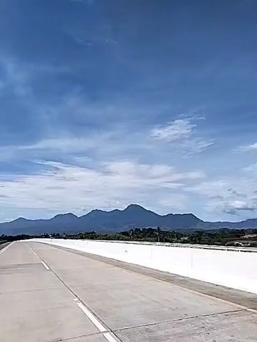 The video shows a road trip on a highway under a blue sky with scattered clouds. The road is wide and concrete, with a white barrier on the side. In the distance, there are mountains covered in greenery. A truck drives along the road.