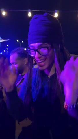 A person with dreadlocks, glasses, and a black head wrap is dancing at an outdoor event. They wear a black jacket and are smiling. The lighting is blue and purple, with string lights overhead. Other people are visible in the background, and music is playing. The person's face is highlighted by colorful lighting effects.