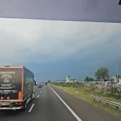 The video shows a view from inside a moving vehicle, likely a truck, on a highway. The highway has multiple lanes, with other vehicles, including cars and trucks, visible. On the right side of the highway, there is a grassy embankment, followed by a fence, and then a building with a control tower and a parking lot. In the distance, a white airplane is seen flying across the sky, which is partly cloudy. The sky appears to be a mix of blue and dark clouds, suggesting potential for rain. The camera is focused on the road and the sky, with the left side of the frame dominated by the rear of the truck the camera is presumably in, displaying text and graphics.