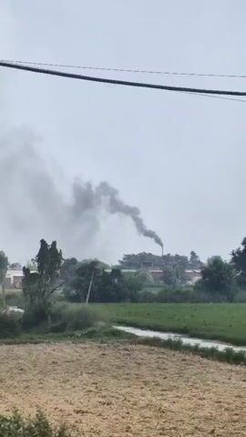 The video begins with a close-up of a red corrugated metal structure, possibly a shipping container. The camera then pans to the left, revealing a wide shot of a rural landscape under a hazy sky. In the distance, a factory chimney is visible, emitting a thick plume of black smoke. The smoke billows and drifts across the sky, obscuring the upper portion of the frame. The surrounding area is dotted with trees and appears to be agricultural land. Electrical wires are also visible in the foreground, with a pole on the left side of the frame. The overall impression is one of industrial activity impacting the natural environment.