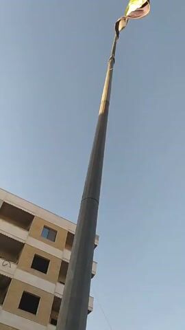 The video shows a tall, gray flagpole standing against a clear blue sky. At the top of the pole, a flag, possibly the flag of Palestine, is waving in the wind. A multi-story building with beige walls and dark windows is visible in the background on the left side of the frame. The camera angle is looking upward towards the flag and flagpole.