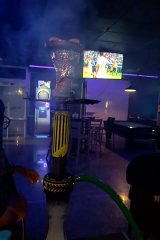 A shisha pipe is in the foreground, with a bright green hose attached to it. In the background, a large screen displays a sporting event, likely soccer, with players on a green field surrounded by spectators in a stadium. The room is dimly lit, with purple and blue accent lights. A few tables and chairs are visible, suggesting a lounge or bar setting. The video is rotated 90 degrees counterclockwise, making it appear vertical.