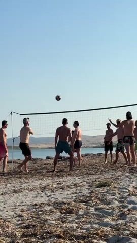 A group of men are playing volleyball on a sandy beach with a clear blue sky overhead. The sun is bright, suggesting it is daytime, likely late afternoon given the angle of the light. The beach is sparsely covered with dried vegetation, and rocks are visible near the water's edge. The water appears calm and blue, hinting at a coastal or lakeside setting. The men are all wearing swim trunks and appear to be in good spirits, engaged in a casual game. Their body language suggests focus and enjoyment of the activity. A volleyball net is set up on the sand, and a volleyball is visible in mid-air, captured during a play. The background shows a slightly hilly, arid landscape under the vast sky. There is no visible text in the frame.