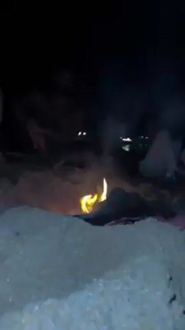 The video depicts a group of people gathered around a bonfire on a beach at night. The fire is the central focus, with flames rising amidst smoke. Individuals are sitting or squatting around the fire; at least three are partially visible. A hand enters the frame, holding a green object that may be a container. The background shows a dark sea, with some lights visible in the distance. The beach sand is light-colored and textured.
