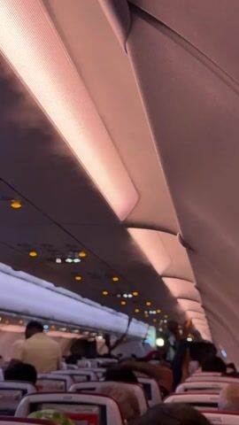 The video shows the interior of an airplane cabin with rows of seats and passengers. The lighting is dim with overhead lights on, casting a pinkish hue. Some passengers are visible in their seats, and others are walking in the aisle. The overhead compartments and part of the airplane's ceiling are also visible. The video appears to be recorded from a passenger's perspective, looking down the aisle of the plane.