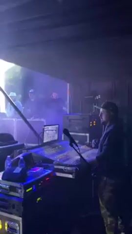 Here is a description of the video.

The video shows a concert or stage performance. In the first frame, a man in a baseball cap and camouflage pants is working at a soundboard. Behind him, people are visible on the stage through a haze or fog. The second frame shows the stage area with people in front of a screen displaying an image of a young person. In the third frame, a crowd of people are watching the stage, which is lit with blue and white lights. The people on stage appear to be setting up or performing. There are also metal support structures visible on the sides of the stage.