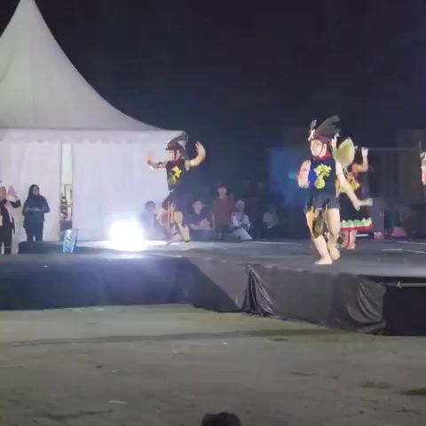 A group of dancers wearing traditional costumes perform on a stage at night. The dancers have elaborate headdresses and are dressed in dark tops and skirts with colorful trim. The stage is lit by bright lights, and a large tent is visible in the background. An audience is seated behind the stage, and a large screen displaying colorful graphics is to the right. The dancers are moving in unison, with some of them holding round objects. The overall atmosphere appears to be a cultural performance.