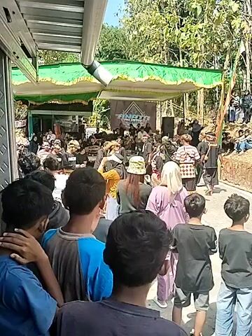 The video shows an outdoor gathering with a stage set up under a green and yellow awning. A group of people, many dressed in traditional attire including headscarves and sarongs, are gathered on and around the stage. Some are seated, while others appear to be performing or interacting. In front of the stage, a crowd of onlookers, primarily young people, are watching the event. The environment appears to be rural, with trees and foliage visible in the background. The overall atmosphere suggests a cultural performance or community event.