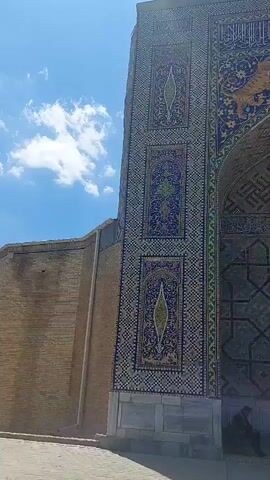 The video shows a large, ornate structure, likely an entrance to a historical site or building, with elaborate mosaic tilework on the facade. The tiles are predominantly in shades of blue, green, yellow, and beige, forming intricate geometric patterns and floral designs. A large arched entrance is visible, with more mosaic work inside the arch. Above the arch, there are depictions of tigers. To the left of the ornate structure is a simpler brick wall. The sky is blue with scattered white clouds. At the bottom of the structure, on the right side, a person is seated. The camera pans slowly to show more of the mosaic structure and the sky.