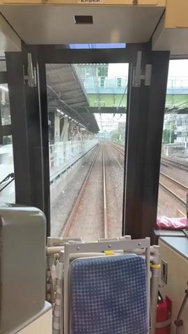 The video shows the view from the front of a moving train. The camera is positioned behind a window, looking forward along the train tracks. To the left is a train station platform with white structures and yellow markings. To the right, there are more train tracks and some buildings. The train appears to be passing through a station or an area with multiple tracks. The sky is overcast. The foreground shows parts of the train's interior, including a seat and some equipment.