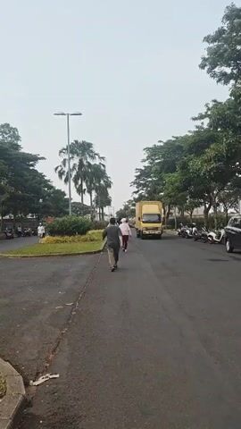 A yellow FUSO truck drives down a paved road. The truck has a white license plate with black text that reads "B2206ITT". The truck's cabin is yellow. The truck bed is also yellow with vertical ridges. The truck has a driver in the cabin. The truck is moving towards the viewer and slightly to the right. On the left side of the road, there is a grass median with several palm trees and a street lamp. There are also some people walking on the sidewalk to the left of the median. On the right side of the road, there are parked motorcycles and a black SUV. The road is lined with trees on both sides. The sky is overcast.