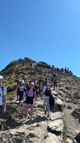 The video begins with a low-angle shot of a crowded, rocky mountain path under a clear blue sky. People, dressed in various casual clothing, are walking up and down the path. The camera then pans right to reveal a train station situated on the mountainside, with a red train parked on the tracks. From there, the camera pans further right to capture a panoramic view of the surrounding mountainous landscape, including lakes and valleys, stretching out towards the sea on the horizon. The sky remains clear and blue throughout. The video ends with a closer view of the mountainous landscape including a lake and the train tracks.