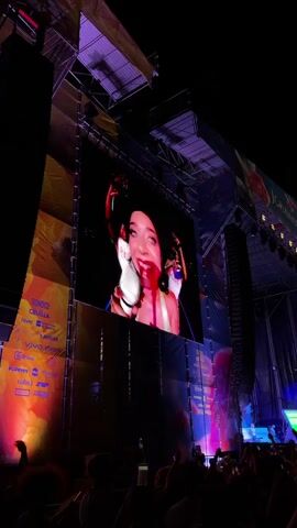 This video captures a night concert. A large screen displays a close-up of a person with their mouth open, appearing to sing or shout, with what looks like fake blood around their mouth. The screen is mounted on a stage structure with lighting equipment and speakers. Below the screen, a crowd of people is visible, many holding up their phones to record or take pictures. The overall environment is dark, illuminated by stage lights and the screen's glow. The crowd is facing the stage, engaged in the performance. The lighting appears to be predominantly purple and pink.