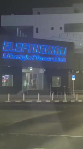 The video shows the exterior of a building at night. The building has a large sign that reads "ELEFTHERIOU Lifestyle Fitness Club". The sign is illuminated with neon lights that change color from blue to green. The building has a black facade with large glass windows. There are stairs leading up to the entrance, which is also illuminated. The street in front of the building is paved and there are white bollards lining the sidewalk. The sky is dark, indicating it is nighttime.
