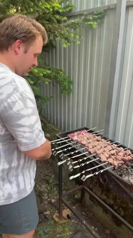 A man in a tie-dye t-shirt and grey shorts is standing next to a grill. He is holding skewers of meat over hot coals. The meat appears to be cubed and is cooking on metal skewers. The grill is black and appears to be made of metal. There is a grey corrugated metal fence behind the grill and some green foliage to the left. The ground is covered in dirt and some grass. Smoke is rising from the grill. The video zooms in on the meat as it cooks.