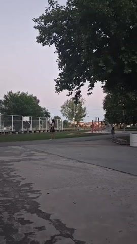 A person stands in a park, holding a phone and looking at it. They are wearing a dark top and shorts. The person is near a fenced-in area, possibly a basketball court. There are trees and playground equipment in the background. The sky is overcast and it appears to be dusk.