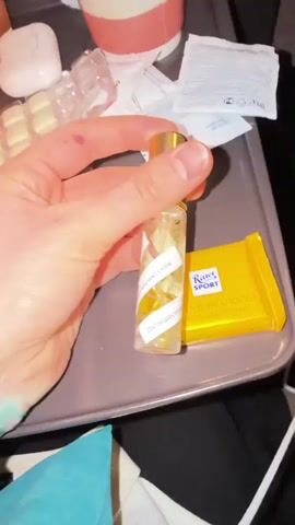 A hand holds a small, clear perfume bottle with a gold top. The bottle has a white label spiraling around it with black text in Russian. The text appears to change as the bottle is rotated in the hand. Inside the bottle, there are what appear to be tiny dried flowers. Next to the bottle is a yellow-wrapped Ritter Sport chocolate bar. The items are resting on a gray tray. Other items are visible in the background, including a blister pack of pills, a pink tube, and a white packet. The background is slightly out of focus.