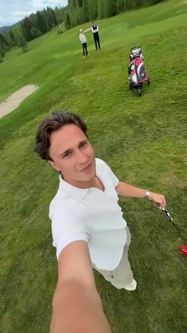 The video shows a person standing on a green grassy area, likely a golf course, holding a golf club. They point the club towards a small, red, square-shaped object on the ground. The person is wearing a white shirt, khaki pants, and white shoes, and they also have a watch on their left wrist. In the background, a sand trap and a hill are visible. The video is shot from a first-person perspective.