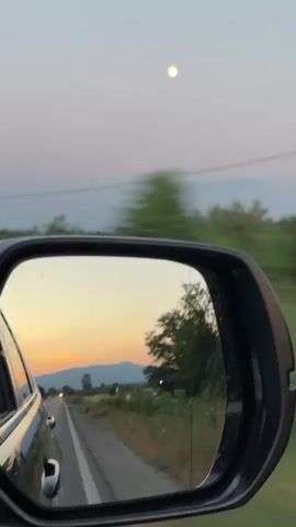 The video shows a view from the passenger side mirror of a car driving down a rural road at dusk. The sky is a pale pink and blue, with a bright, hazy orb, likely the moon, visible in the upper portion of the frame. The mirror reflects the road ahead, with the car's side visible on the left. The landscape outside the car appears to be agricultural, with fields and trees lining the road. There are distant hills or mountains visible on the horizon. The time of day is clearly sunset or twilight, indicated by the soft light and pastel colors in the sky and reflection. The activity is a car journey through a scenic, possibly countryside setting. There are no people visible, only the car and the landscape. No text is visible in the video. The overall mood is calm and contemplative, with the motion blur of passing trees adding a sense of movement and transience.