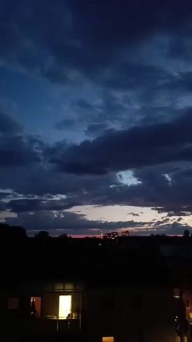 The video captures a static, vertical shot of a dark evening sky filled with low, dark blue clouds. A narrow band of orange and red light is visible on the horizon, beneath which a dark silhouette of a cityscape and trees stretches across the frame. In the lower left foreground, the top of a dark building is visible, with one window illuminated by a warm yellow light.