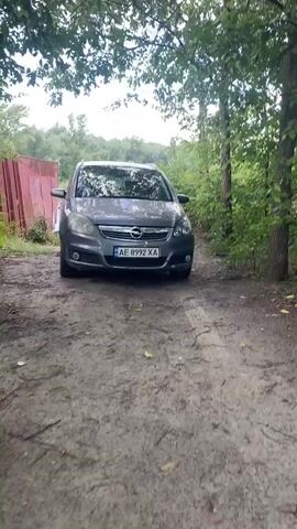 A gray Opel Zafira is parked on a dirt road. To the left of the car, there is a red metal structure. To the right of the car, there are trees. A person walks from behind the red structure and stands near the front of the car.