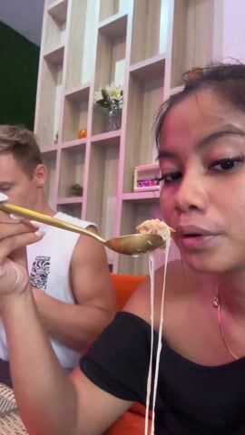A woman with long eyelashes and a black off-the-shoulder shirt is sitting in a restaurant, holding a spoonful of cheesy food up to her mouth, then putting it down and looking at something in front of her. The woman's hair is pulled back in a loose bun. She is wearing a gold necklace. In the background, a man in a white tank top is looking down, presumably eating. Behind them is a decorative shelf with plants, vases and small decorative objects. The restaurant is illuminated with pink and white lighting.